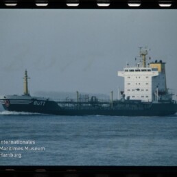 Tanker mit weißen Aufbauten und dunkelblauem Rumpf mit dem Namen BUTT fährt über ruhige See; Museumslogo unten links.