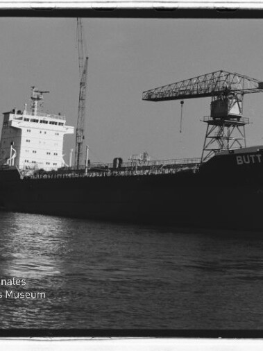 Frachtschiff BUTT liegt im Hafen neben einem großen Kran, schwarzweißes Foto mit Wasser im Vordergrund.