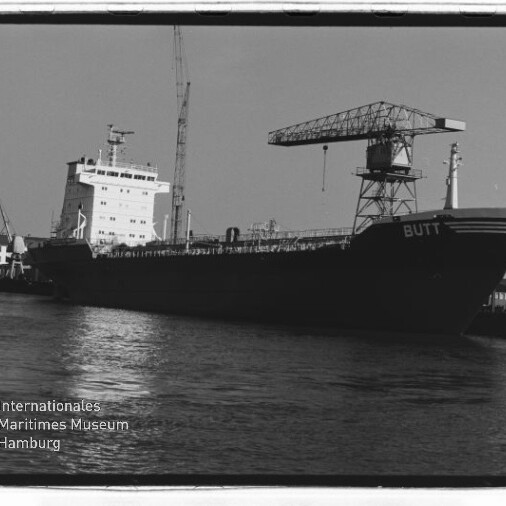 Frachtschiff BUTT liegt im Hafen neben einem großen Kran, schwarzweißes Foto mit Wasser im Vordergrund.
