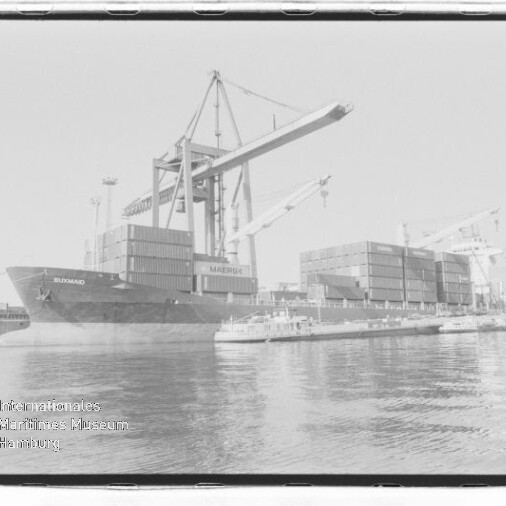 Großes Containerschiff am Kai mit Hafenkränen, gestapelten Containerblöcken und einem kleinen Schubboot im Vordergrund.