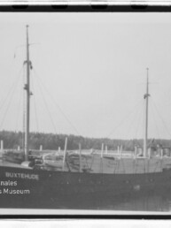 Zweismastiger Frachter BUXTEHUDE vor bewaldeter Küste auf ruhigem Wasser, schwarzweiß.