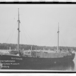 Zweismastiger Frachter BUXTEHUDE vor bewaldeter Küste auf ruhigem Wasser, schwarzweiß.