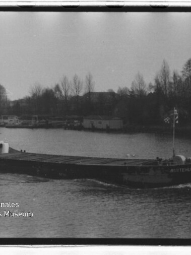 Ein langes Frachtschiff fährt auf einem Fluss, im Hintergrund Ufer mit kahlen Bäumen und Häusern; Museumssignet unten links.