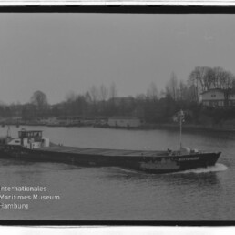 Binnenschiff mit niedrigem Aufbau fährt auf einem Fluss, im Hintergrund Ufer mit Häusern und kahlen Bäumen bei trübem Himmel.