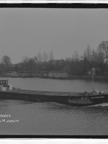 Binnenschiff mit niedrigem Aufbau fährt auf einem Fluss, im Hintergrund Ufer mit Häusern und kahlen Bäumen bei trübem Himmel.