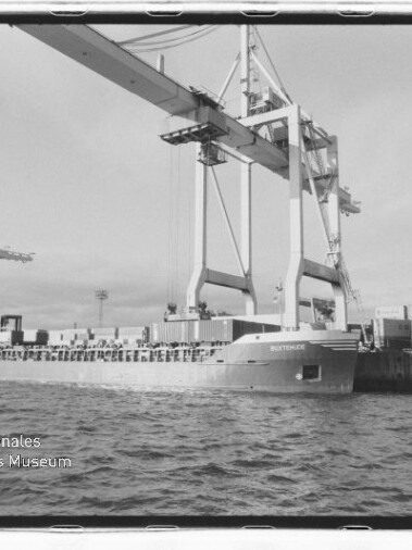 Containerschiff am Kai unter zwei riesigen Hafenkranen, Containerstapel am Pier und Wasser im Vordergrund.