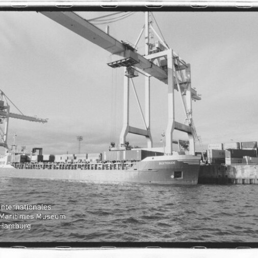 Containerschiff am Kai unter zwei riesigen Hafenkranen, Containerstapel am Pier und Wasser im Vordergrund.