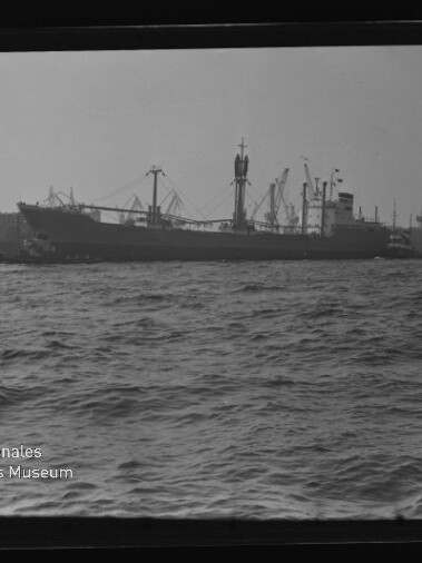Frachtschiff am Hafen mit Kränen und welliger See im Vordergrund; sichtbares Wasserzeichen 'Internationales Maritimes Museum'.