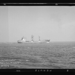 Schwarzweißaufnahme eines Frachtschiffs mit Ladekränen auf ruhiger See am Horizont.