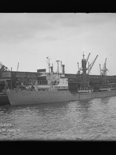 Frachtschiff liegt am Industriehafen vor Lagerhallen, zahlreiche Hafenkräne ragen über ruhiges Wasser.