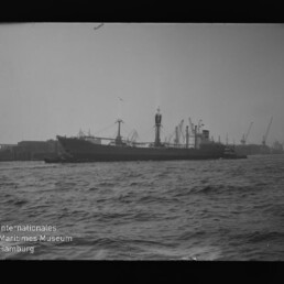 Schwarzweißfoto eines Frachtschiffs im Hafen mit Kränen im Hintergrund und Wellen im Vordergrund; Museumssignet links unten.