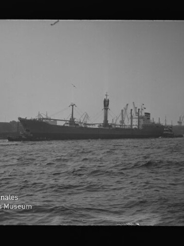 Schwarzweißfoto eines Frachtschiffs im Hafen mit Kränen im Hintergrund und Wellen im Vordergrund; Museumssignet links unten.