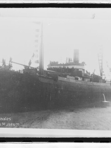 Schwarzweißfoto eines Dampfschiffs am Kai, Personen auf dem Deck und Museumsemblem links unten.