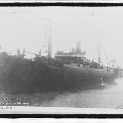 Schwarzweißfoto eines Dampfschiffs am Kai, Personen auf dem Deck und Museumsemblem links unten.