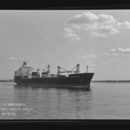 Frachtschiff mit Ladekränen auf ruhigem Wasser und leicht bewölktem Himmel; links unten Schriftzug Maritimes Museum Hamburg