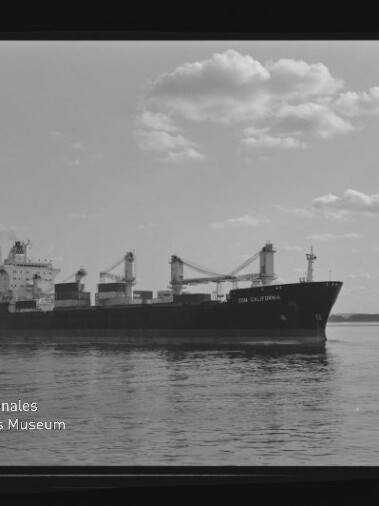 Frachtschiff mit Ladekränen auf ruhigem Wasser unter bewölktem Himmel.
