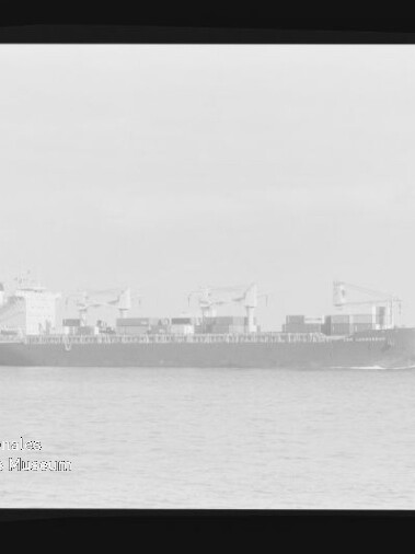 Schwarzweißfoto eines Containerschiffs mit Ladekränen und gestapelten Containern auf ruhiger See, Museumswasserzeichen unten links.