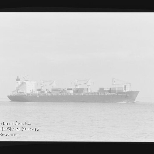 Schwarzweißfoto eines Containerschiffs mit Ladekränen und gestapelten Containern auf ruhiger See, Museumswasserzeichen unten links.