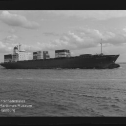 Containerschiff mit geladenen Containern fährt bei bewölktem Himmel; Schriftzug des Maritimen Museums Hamburg sichtbar.