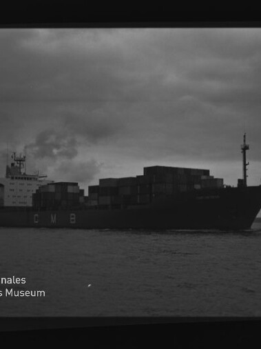 Containerschiff mit aufgestapelten Containerkästen und CMB-Schrift auf dem Rumpf fährt bei dunklem, bewölktem Himmel auf dem Meer; Museumslogo und Filmrand sichtbar.