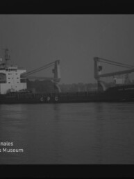 Frachtschiff mit zwei Ladekränen und 'CPC' am Rumpf auf ruhigem Wasser vor Hafenkulisse; Museumslogo links.