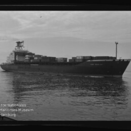Containerschiff mit aufgestapelten Containern auf ruhiger See, Seitenansicht; Museumslogo unten links