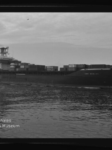 Containerschiff mit aufgestapelten Containern auf ruhiger See, Seitenansicht; Museumslogo unten links