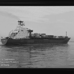 Schwarzweißaufnahme eines Containerschiffs mit gestapelten Containern auf ruhigem Wasser, Museumslogo unten links.