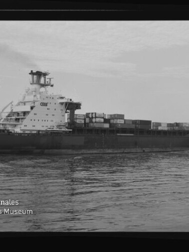 Schwarzweißaufnahme eines Containerschiffs mit gestapelten Containern auf ruhigem Wasser, Museumslogo unten links.