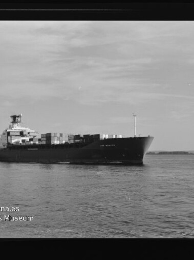 Containerschiff mit aufgestapelten Containern auf ruhiger See vor flacher Küste, Museumslogo links unten.