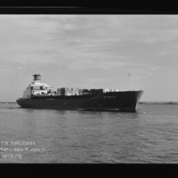 Containerschiff auf ruhigem Wasser, seitliche Ansicht mit Containern; Wasserzeichen: Internationales Maritimes Museum.