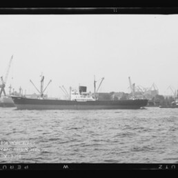 Frachtschiff im Hafen vor Kränen, begleitet von einem kleinen Schlepper, schwarzweißes Bild.