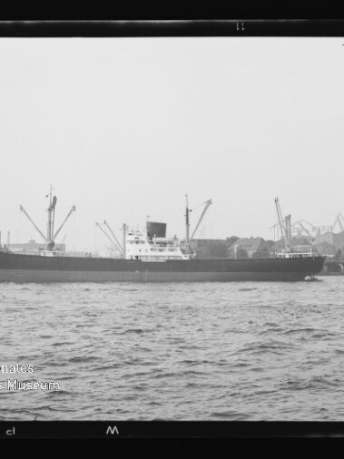 Frachtschiff im Hafen vor Kränen, begleitet von einem kleinen Schlepper, schwarzweißes Bild.