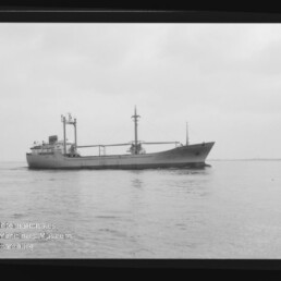 Frachtschiff mit zwei Masten und Ladegeschirr fährt auf ruhigem Wasser; Museumslogo unten links sichtbar.