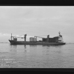 Frachtschiff mit Ladekränen und Containern auf ruhiger See, Museumslogo unten links.