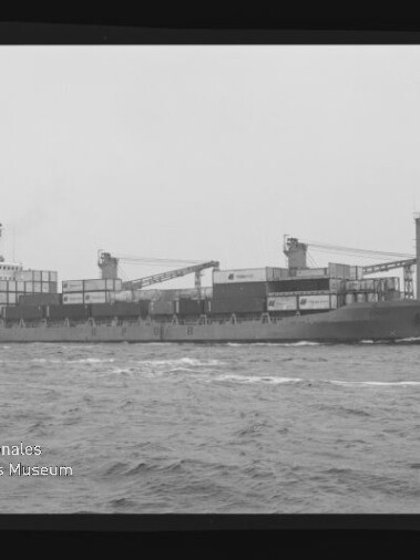 Frachtschiff mit gestapelten Containern und zwei Ladekränen auf ruhiger See, schwarz-weißes Foto.