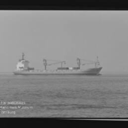 Schwarzweißfoto eines Frachtschiffs mit Ladekränen auf ruhiger See; Filmrand und Beschriftung AGFA PAN 100 sichtbar.