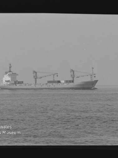 Schwarzweißaufnahme eines Frachtschiffs mit zwei Ladekranen auf ruhiger See, Logo Internationales Maritimes Museum Hamburg unten links.