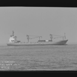 Frachtschiff mit zwei Ladekränen auf ruhiger See, schwarzweißes Foto mit Wasserzeichen Internationales Maritimes Museum Hamburg
