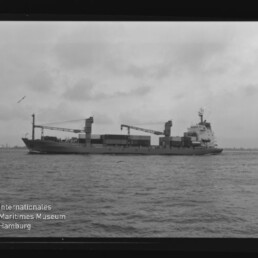 Schwarzweißfoto eines Frachtschiffs mit Ladekränen auf ruhiger See; Stempel Internationales Maritimes Museum Hamburg.