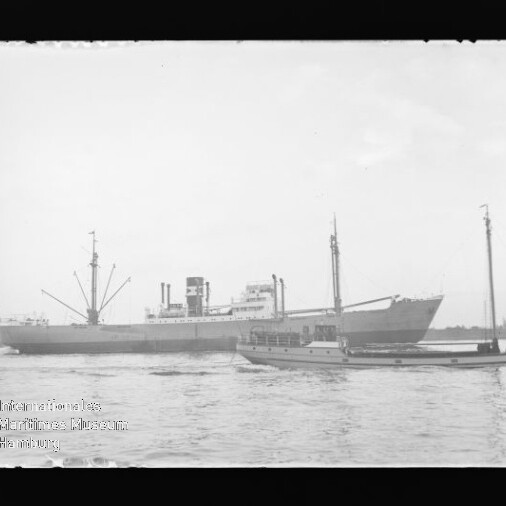 Großes Frachtschiff mit Ladebäumen und Schornstein, begleitet von einem kleineren Boot auf ruhigem Wasser vor flachem Ufer