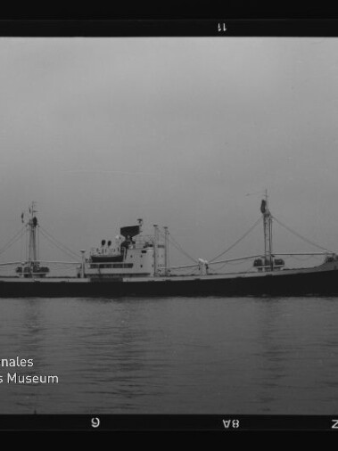 Schwarzweißfoto eines Frachters mit zwei Ladebäumen auf ruhiger See, Museumssignet Internationales Maritimes Museum Hamburg links unten.