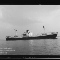 Schwarzweißaufnahme eines Frachtschiffs auf ruhiger See mit sichtbaren Ladebäumen und Aufbauten, Wasserzeichen Internationales Maritimes Museum Hamburg unten links.