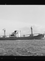 Frachtschiff in Seitenansicht auf ruhiger See mit Mast und Ladebaum, Museumslogo unten links.