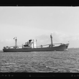 Frachtschiff in Seitenansicht auf ruhiger See mit Mast und Ladebaum, Museumslogo unten links.