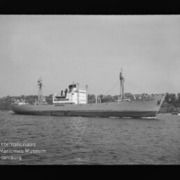 Frachtschiff mit Aufbauten und Ladekränen auf ruhigem Wasser, Baumreihe am Ufer; Museumslogo unten links sichtbar.