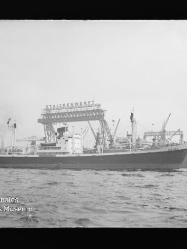 Frachtschiff vor Werft mit Krananlagen und großem Schriftzug, Wellen im Vordergrund.