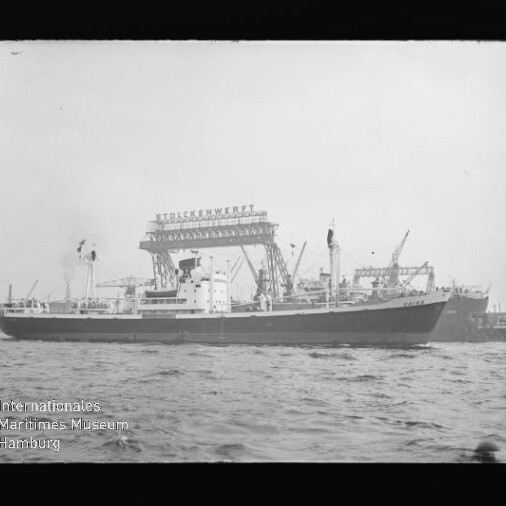 Frachtschiff vor Werft mit Krananlagen und großem Schriftzug, Wellen im Vordergrund.