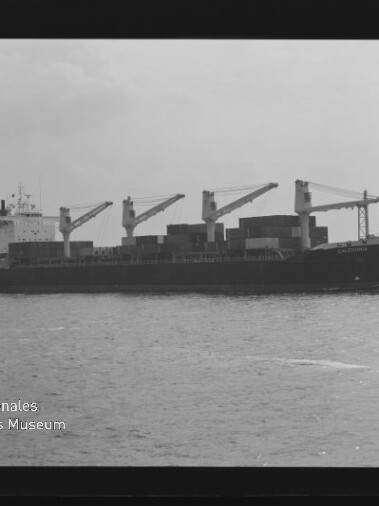 Schwarzweißaufnahme eines Containerschiffs mit vier Ladekränen und gestapelten Containern auf ruhiger See.