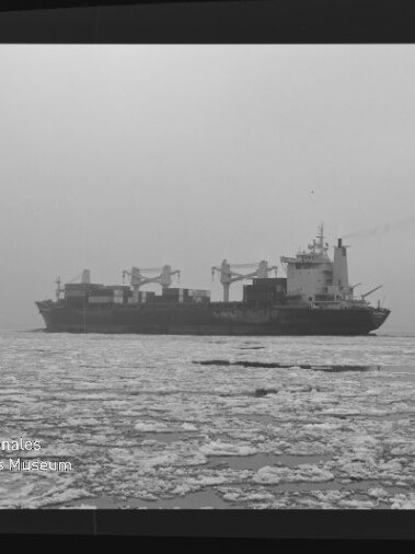Frachtschiff mit Ladekränen fährt durch ein mit Eisschollen bedecktes Meer bei nebligem Himmel.
